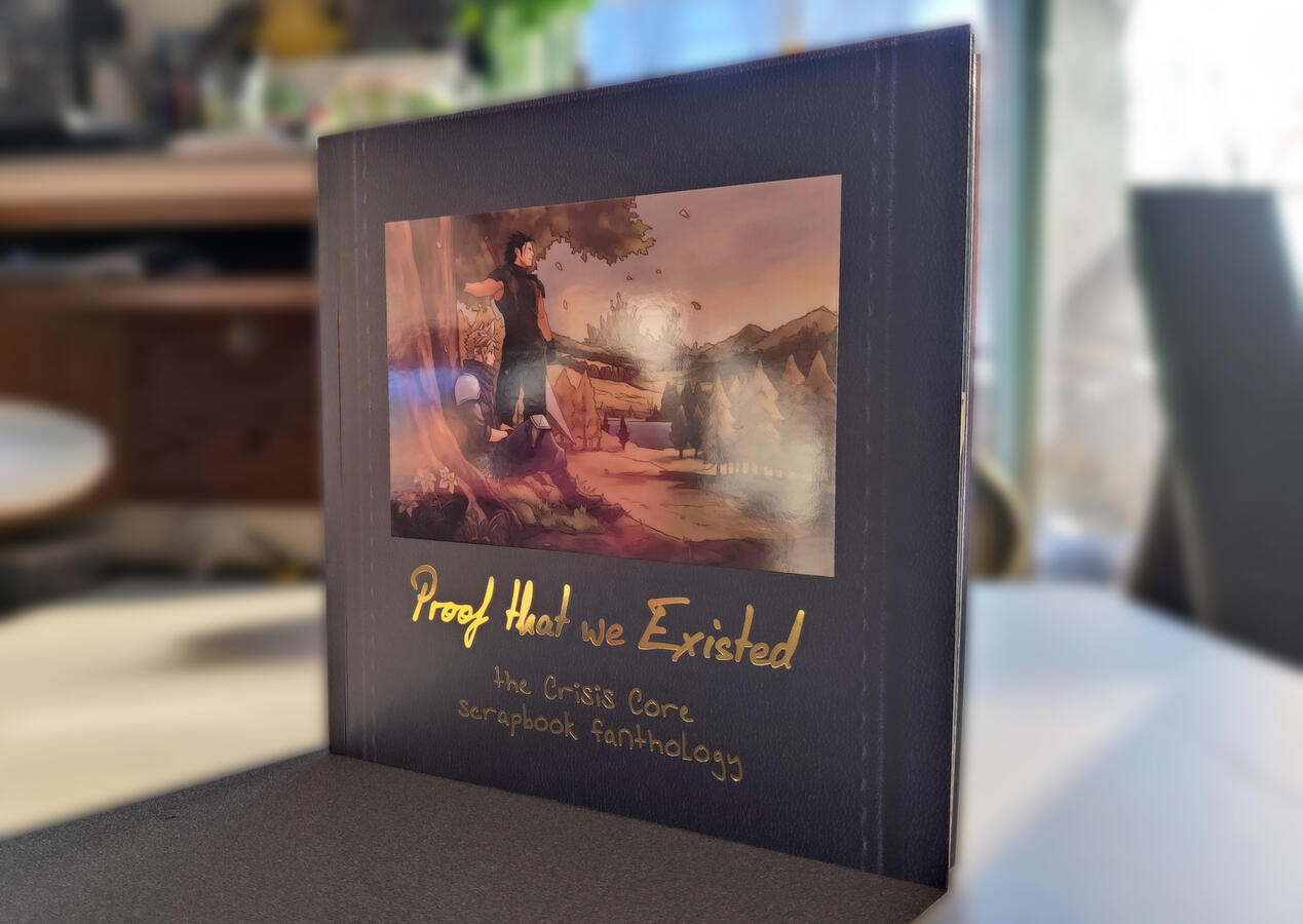 Photo of the front cover of the completed Crisis Core Scrapbook zine. It is facing away from the light source to better show the shine of the Spot UV and gold foil text. The background has been blurred to focus the image.